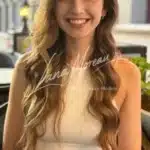 Beautiful companion Kate with long blonde waves and bright brown eyes, smiling sweetly in a sleeveless white top – radiating charm and youth.