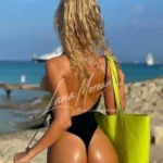 blonde escort in black swimsuit beachside